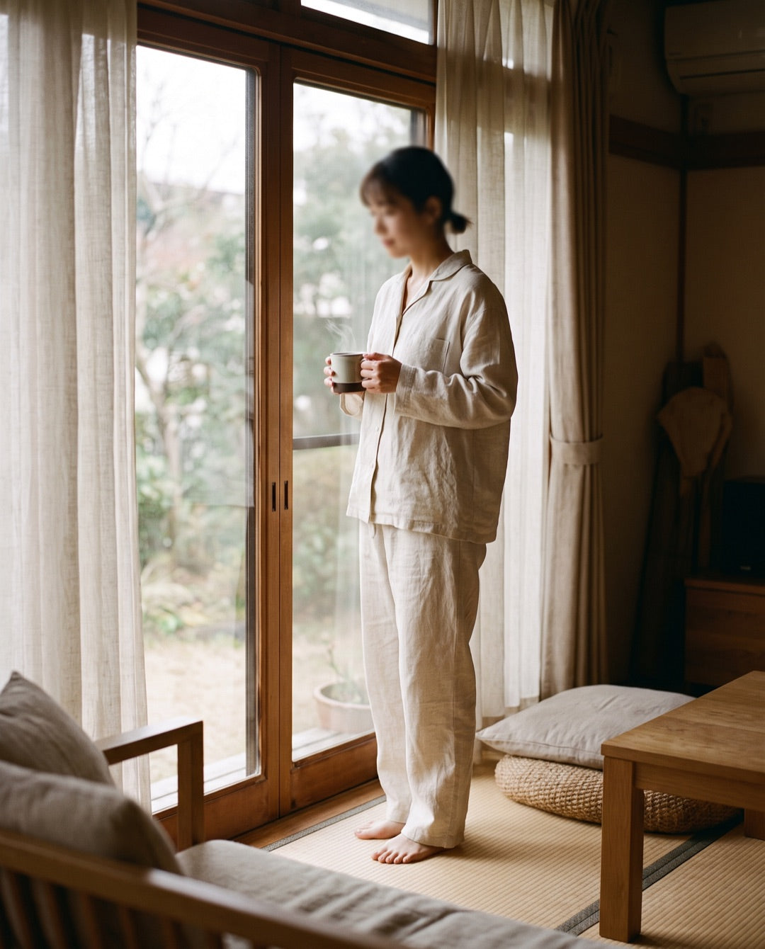 朝の窓辺。淹れたてのコーヒーを手に、自然光の中でくつろぐ、VELUNEのラウンジウェアを着た様子。